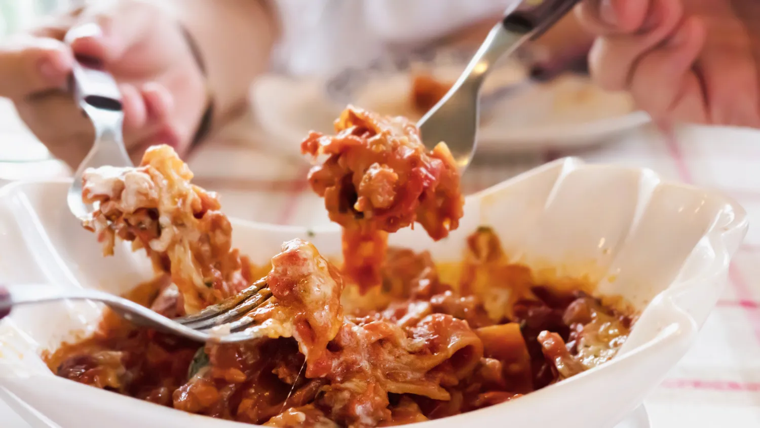 Plato de pasta con salsa de tomate, carne y queso, servido en un tazón blanco, mientras una persona utiliza un tenedor para servir. Fondo desenfocado que sugiere un ambiente acogedor de comida.