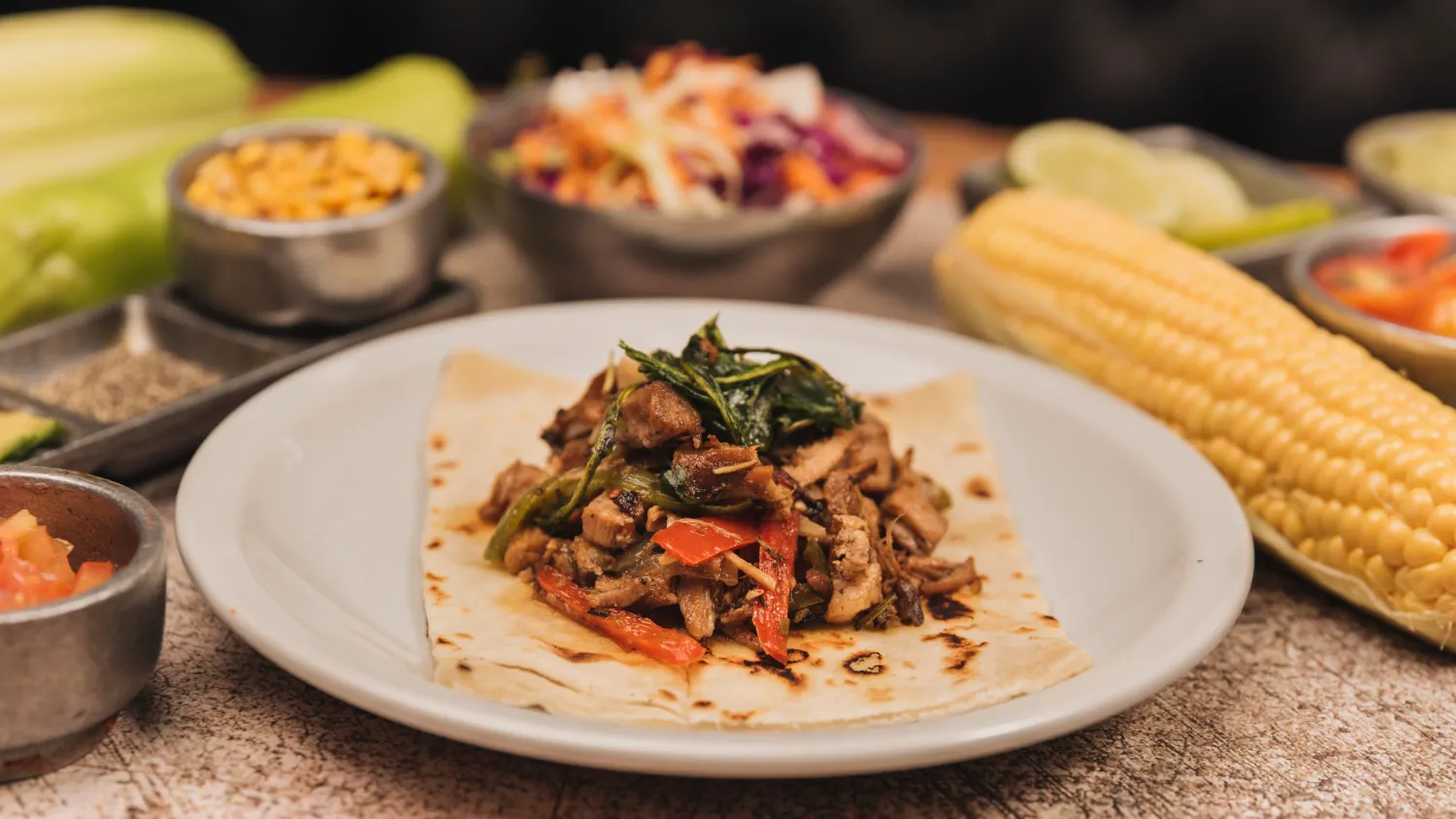 Plato de carne asada servida sobre una tortilla, acompañada de verduras salteadas, maíz, salsa y guarniciones frescas en un entorno acogedor.