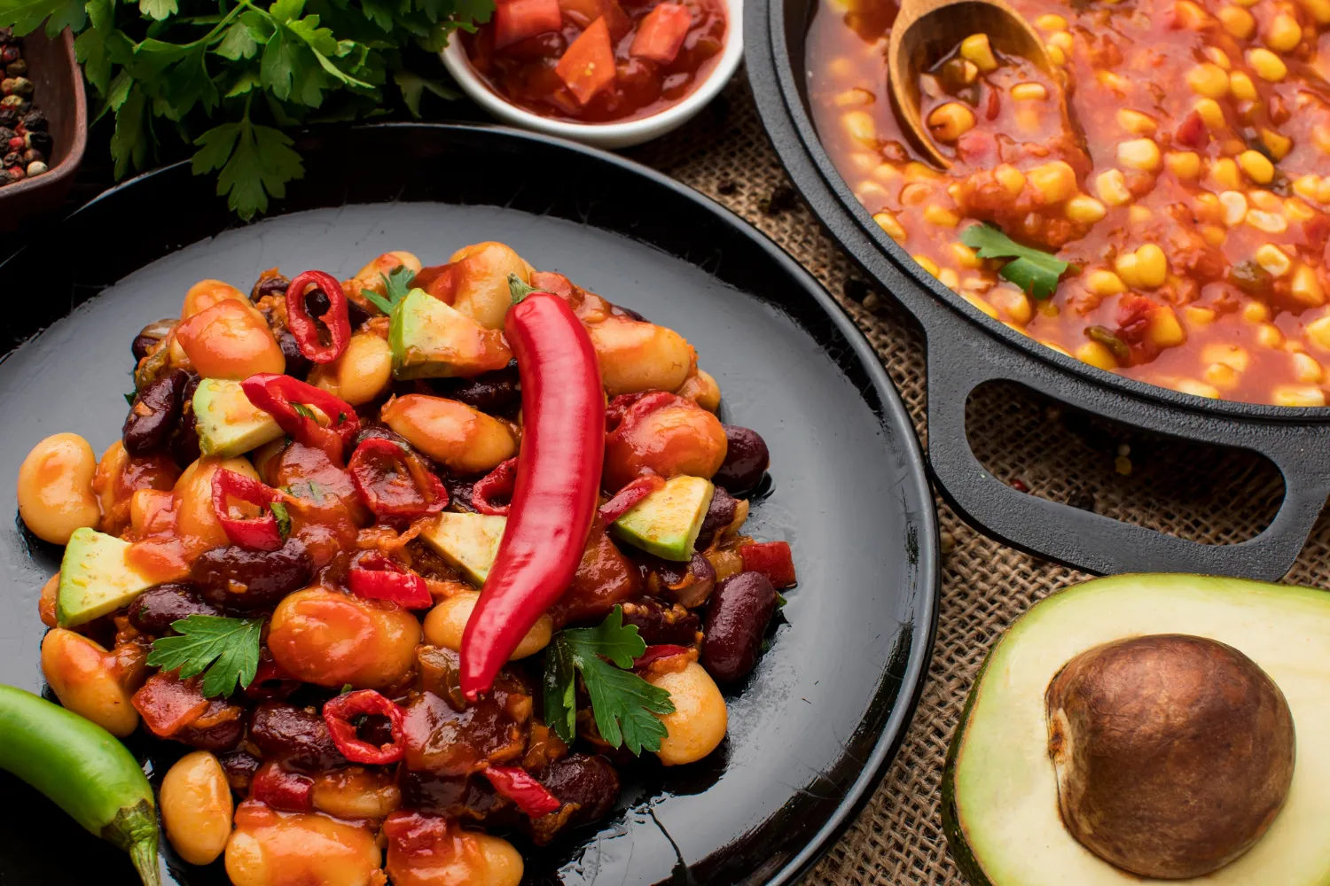 Plato colorido de frijoles variados con pimientos, calabacines y perejil, acompañado de un guiso de maíz y tomate en una sartén. Ideal para una comida saludable y nutritiva.